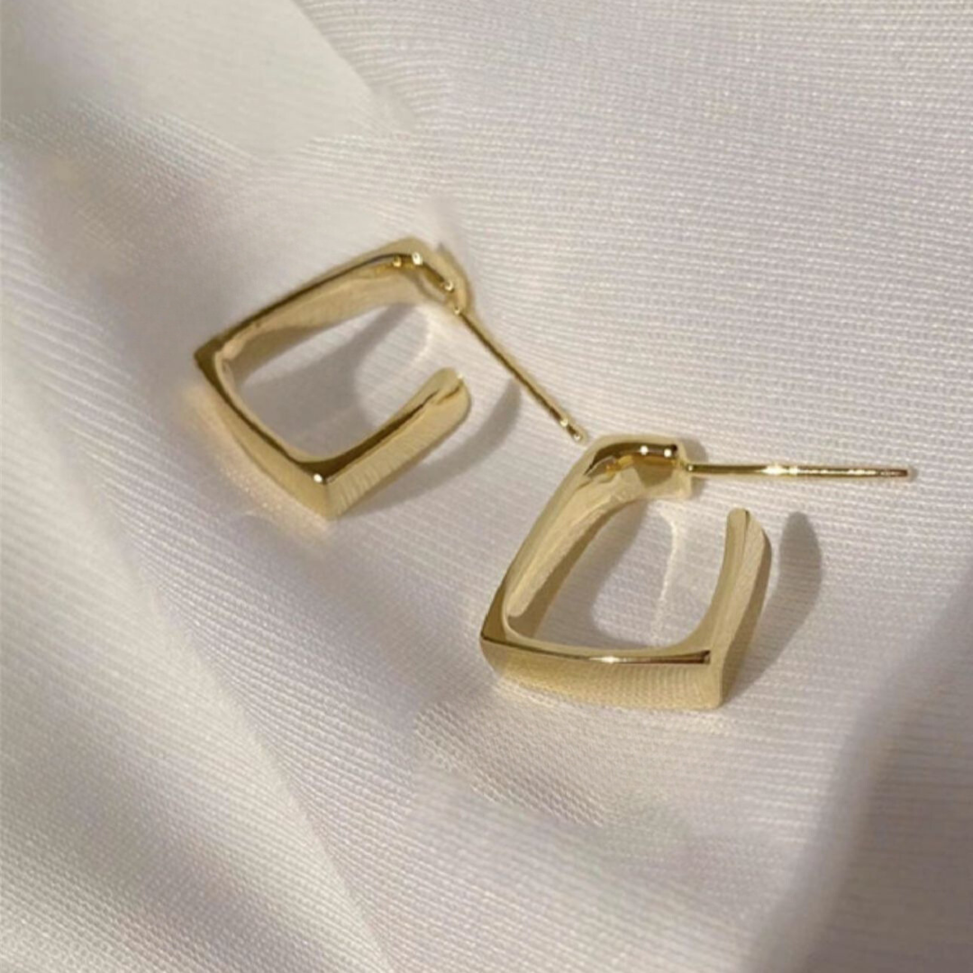 A pair of square-shaped earrings with a gold-tone finish on a white fabric surface.
