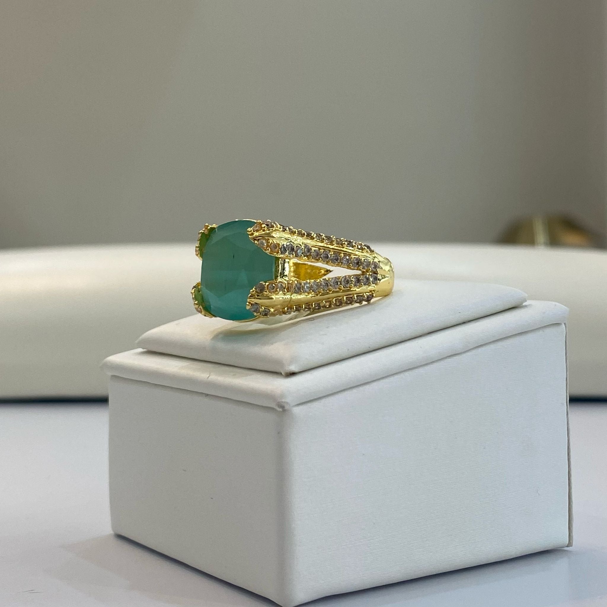 An elegant gold-colored adjustable gemstone ring with a prominent green centerpiece stone, displayed on a white box.