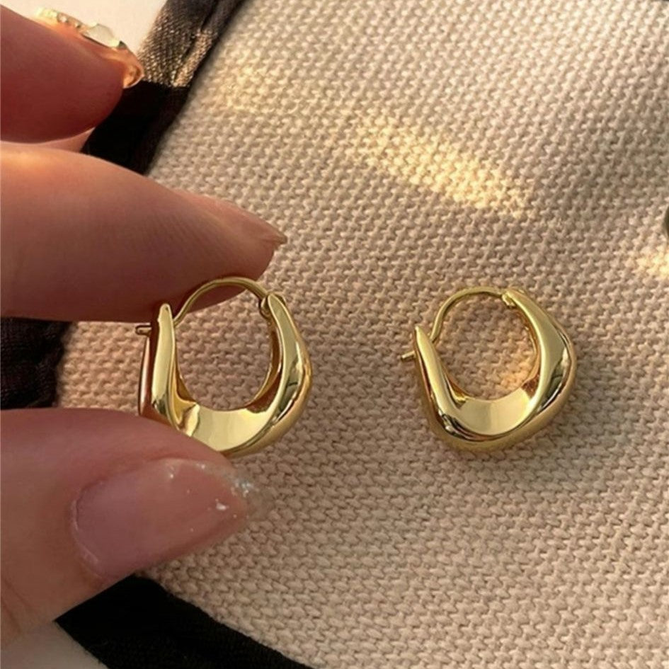 A pair of gold-colored hoop earrings with a curved design, displayed against a beige fabric background.