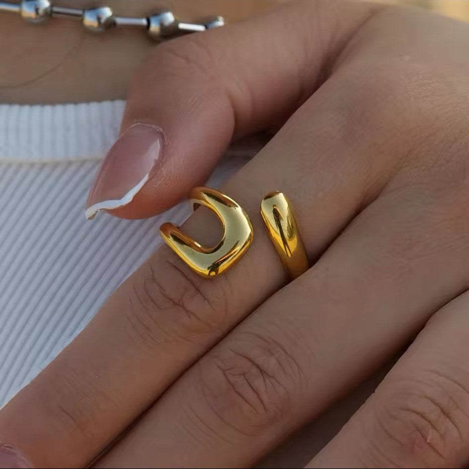 A gold-colored stainless steel hollow layered wide ring is displayed on a female's hand with white surface in the background.