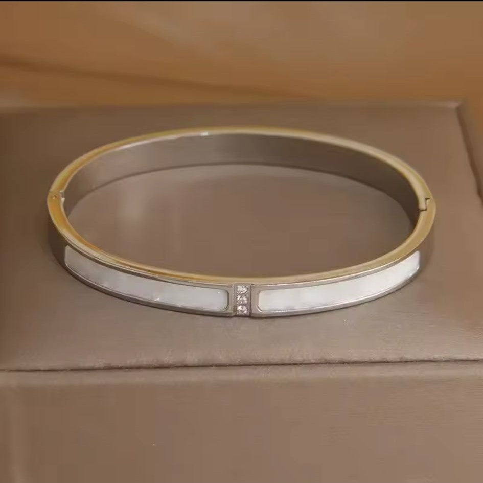 A silver stainless steel bangle bracelet with a pearl shell accent, displayed on a textured surface.