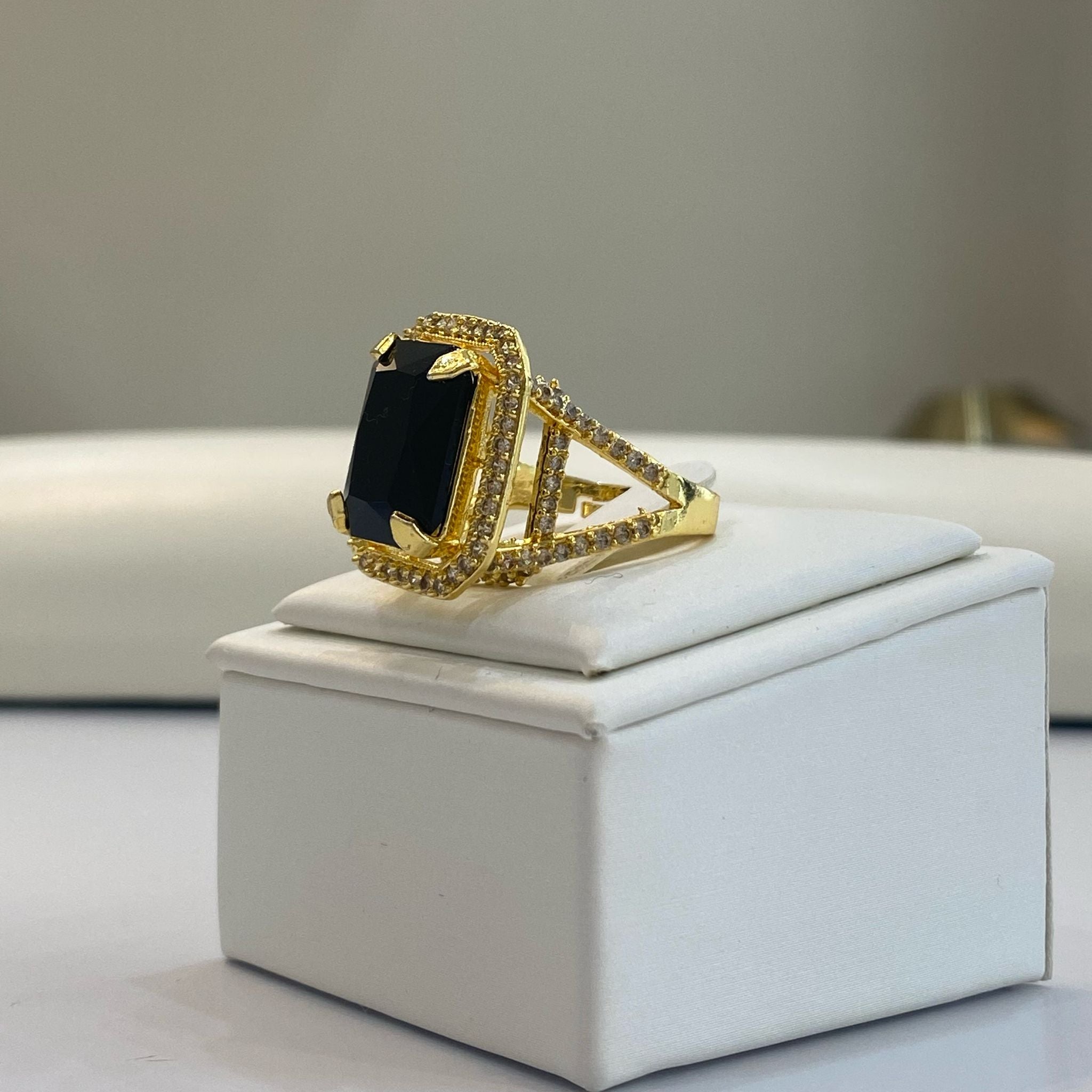 An elegant emerald black gemstone ring with a gold-colored band, featuring intricate detailing, displayed on a white square pedestal.