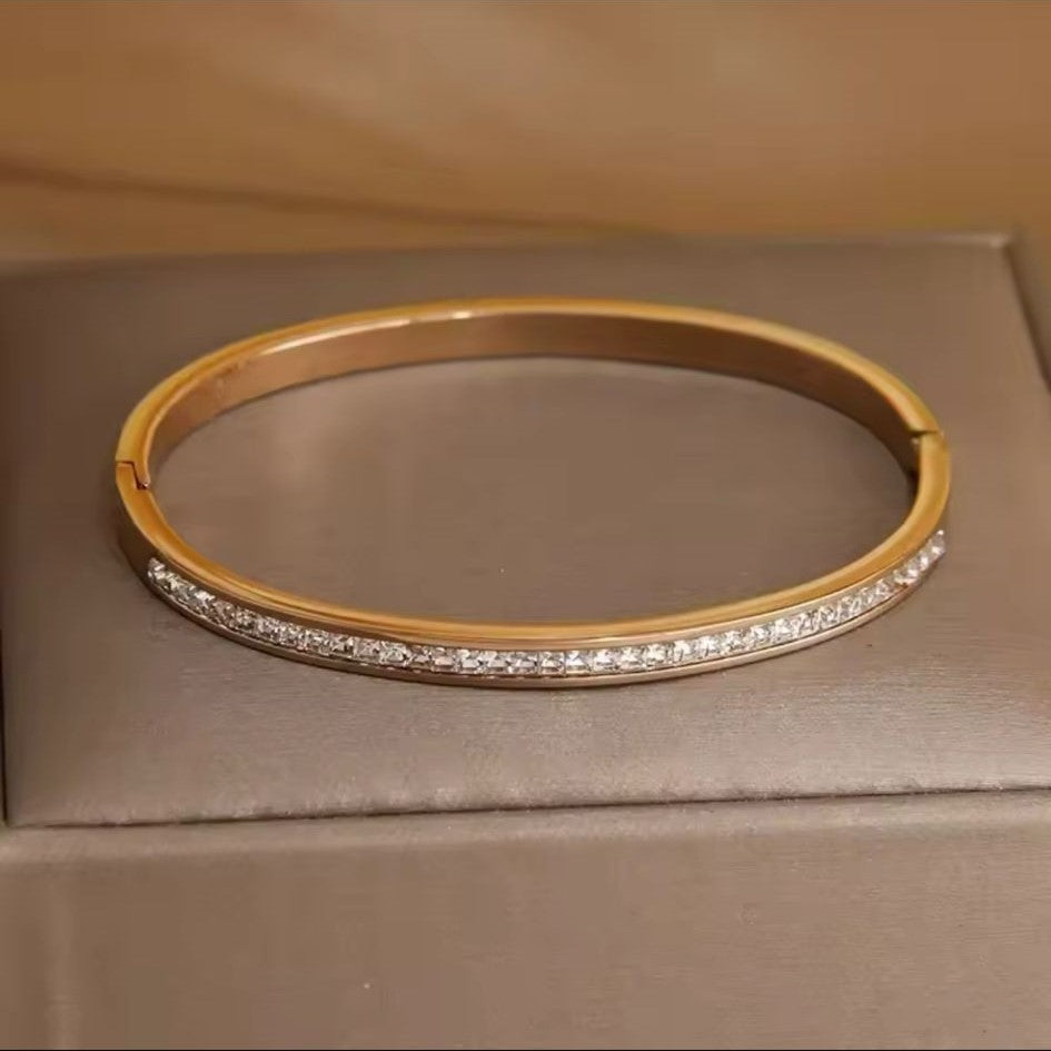 A gold stainless steel bangle bracelet with diamond accents displayed on a textured surface.