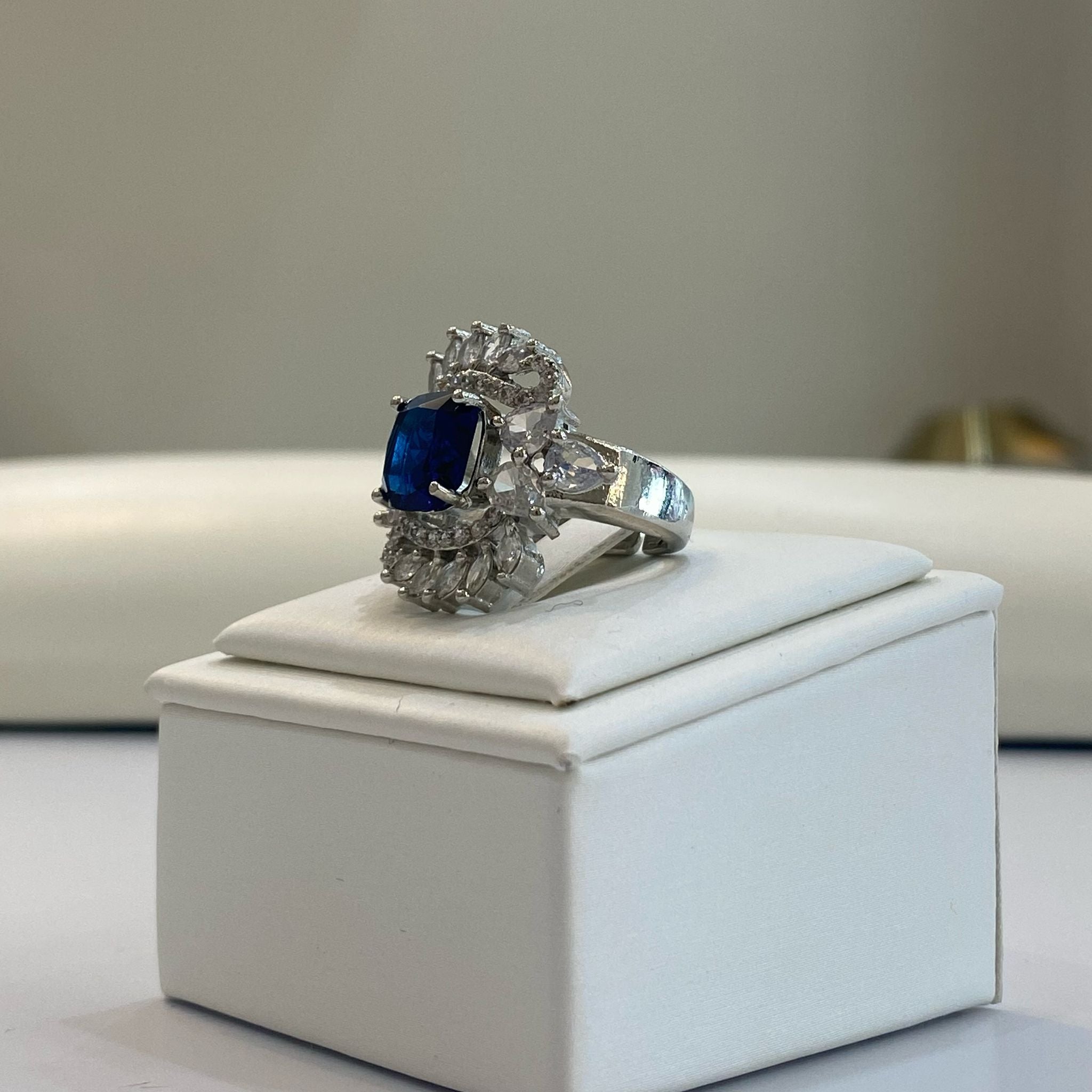An elegant silver adjustable gemstone ring with a prominent blue center stone and intricate detailing, displayed on a white box.