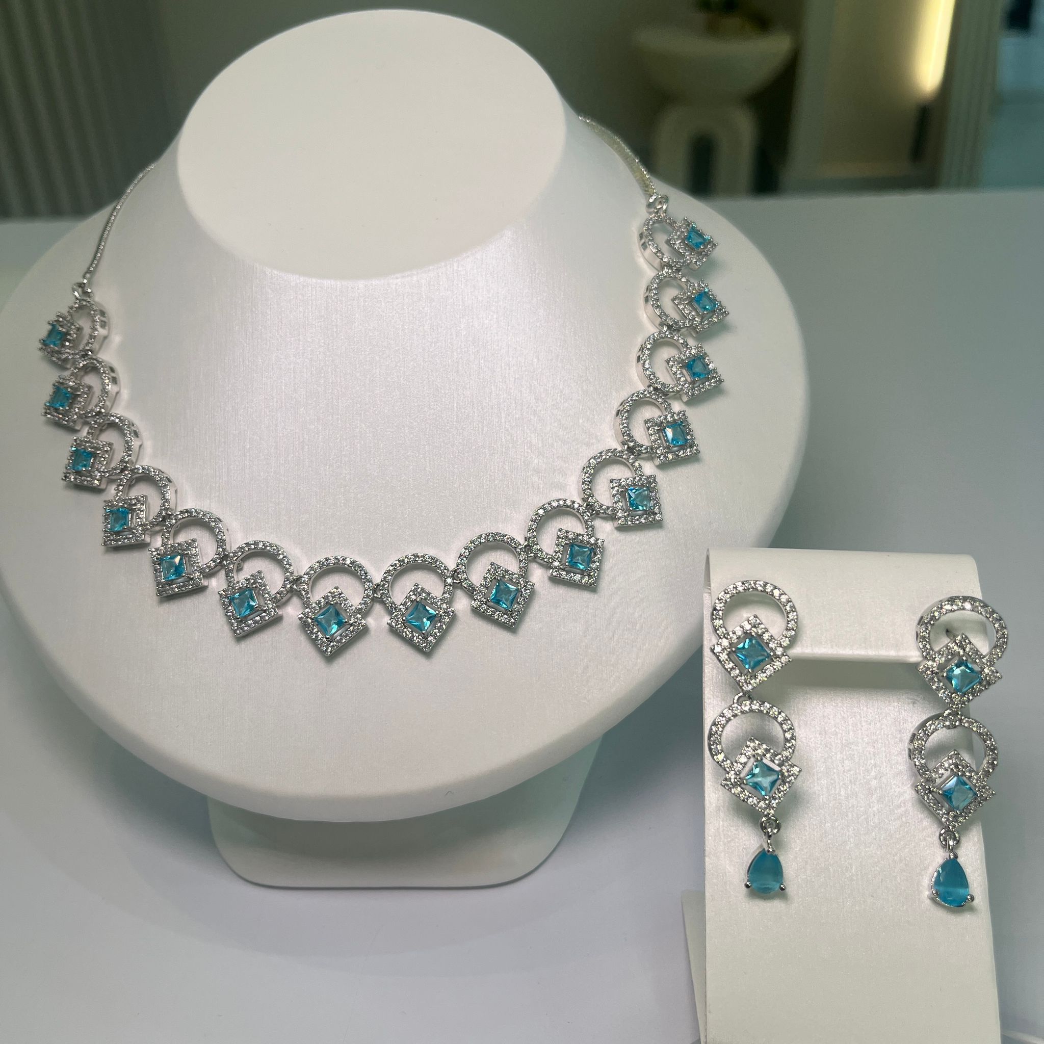 A jewelry set showcasing a wave-like necklace and matching earrings, adorned with blue and clear crystals, displayed on a white mannequin bust.