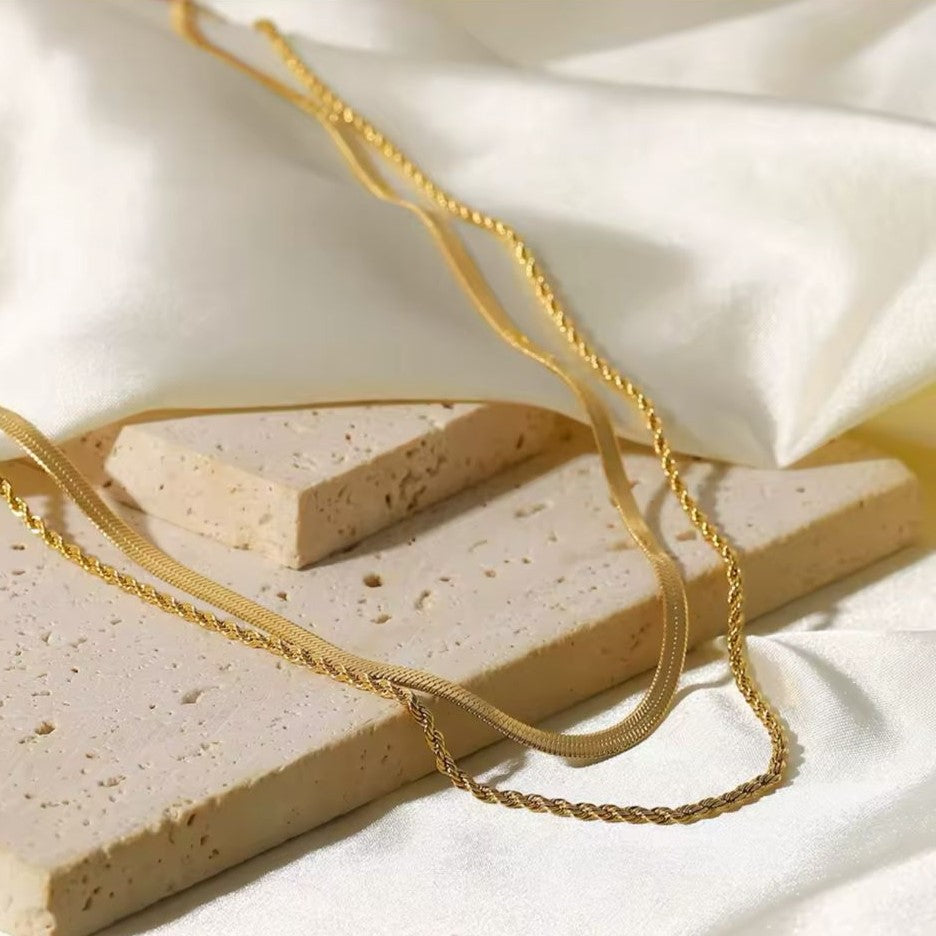A gold colored twin chain necklace displayed on a beige background.