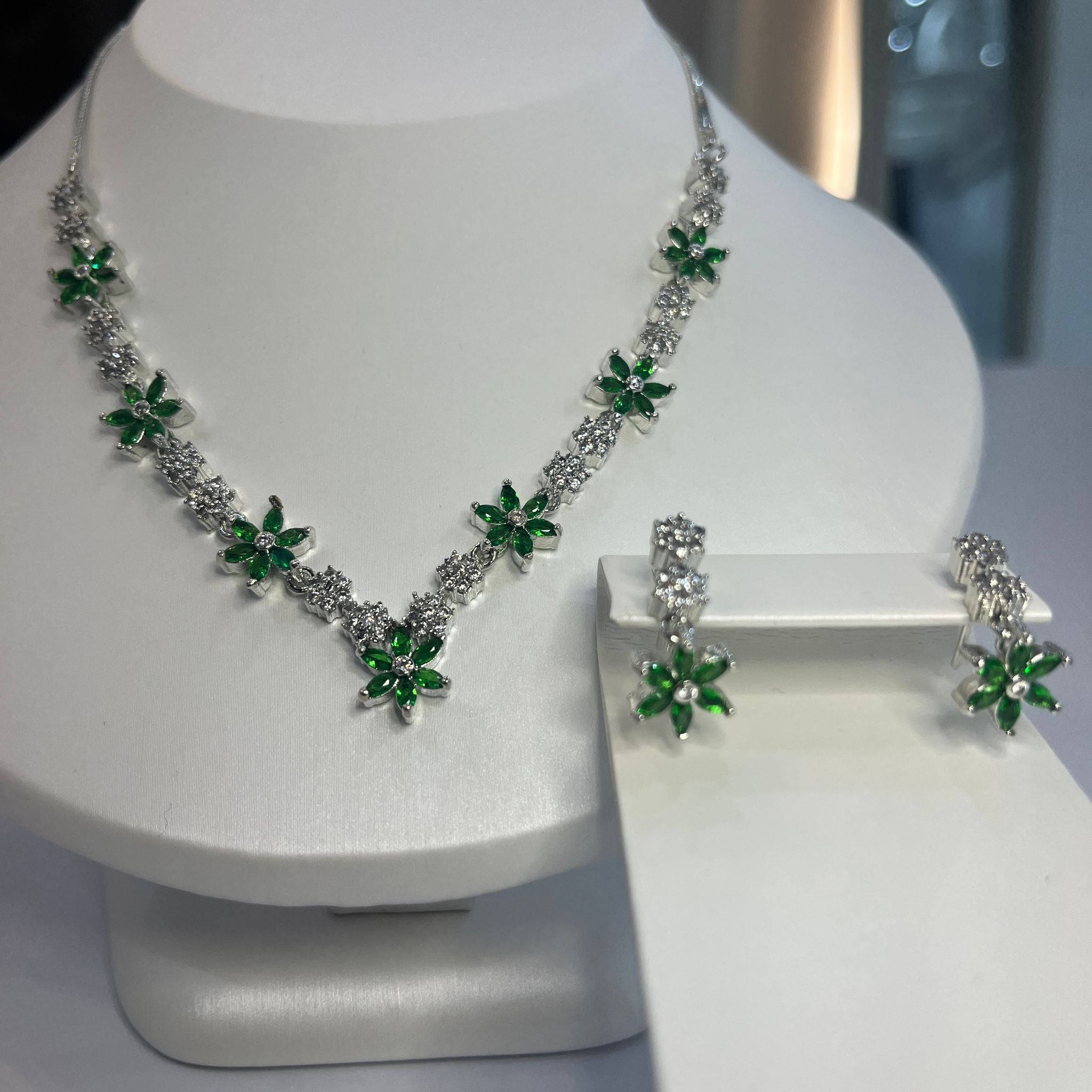 A jewelry set featuring a floral-inspired necklace with green and white stones and matching drop earrings, displayed on a white mannequin.