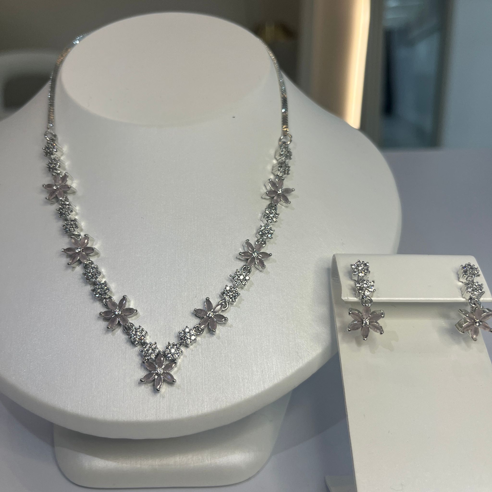 A jewelry set featuring a floral-inspired necklace with clear white stones and matching drop earrings, displayed on a white mannequin.