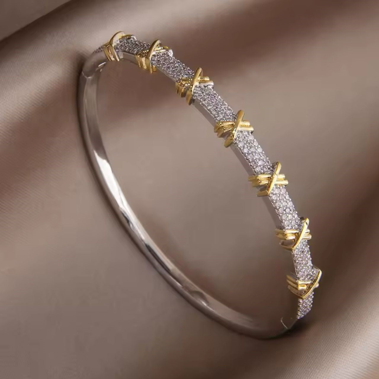 A silver bangle bracelet with gold-tone 'X' accents and sparkling crystal embellishments, displayed on a satin-like surface.
