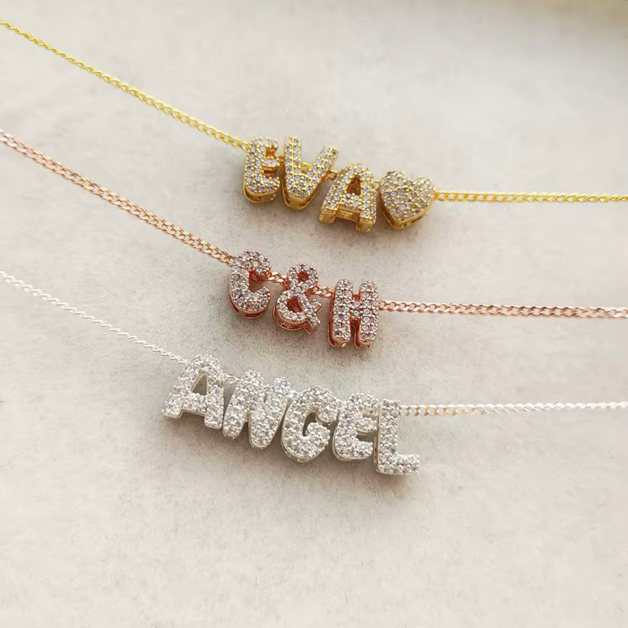 Three personalized bubble letter charm necklaces in gold, silver and bronze tone, with each necklace displaying charms in different sizes and styles, set against a white background.