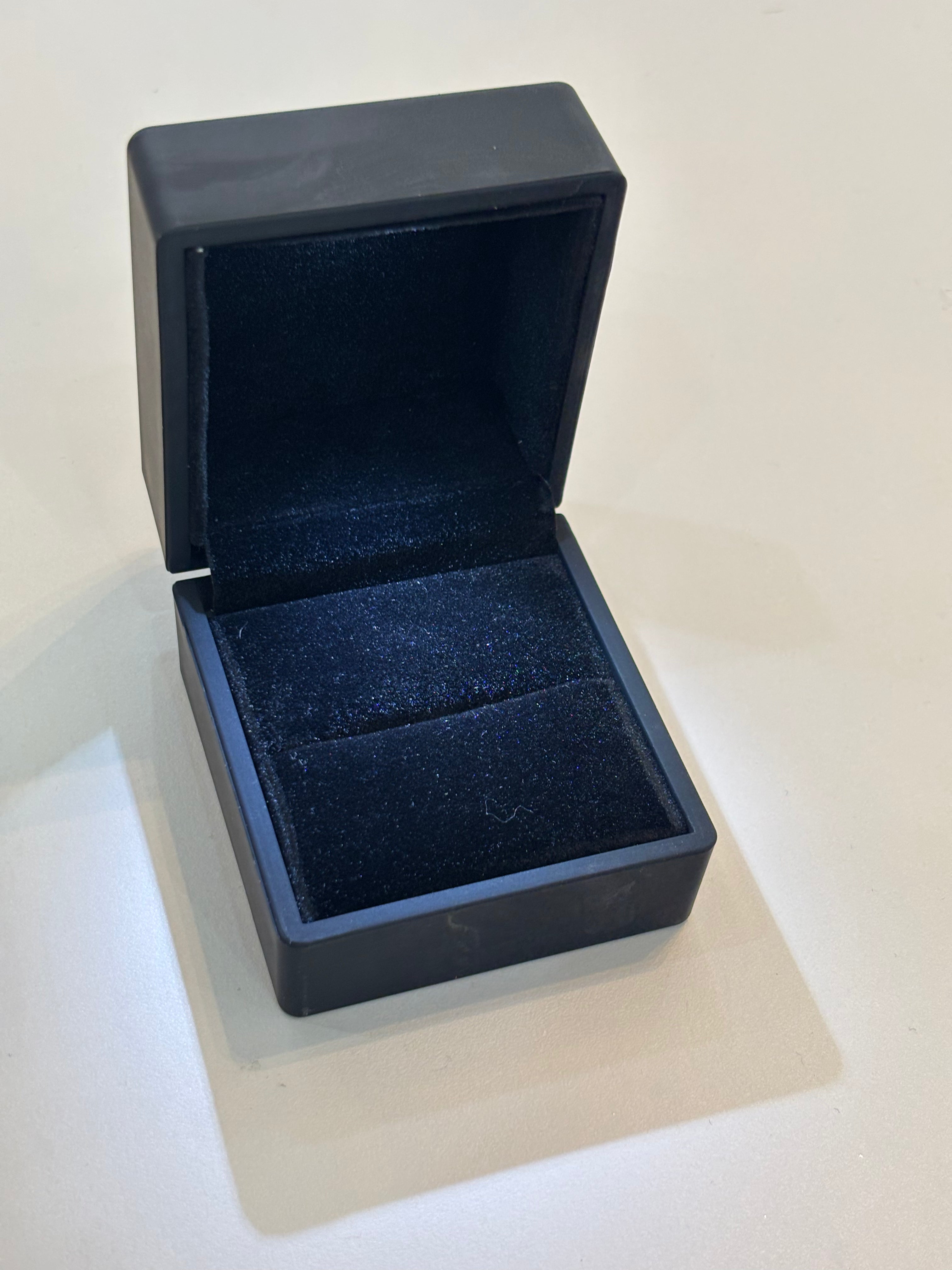 A black ring box with velvet inside. The box is open and positioned on a white surface.