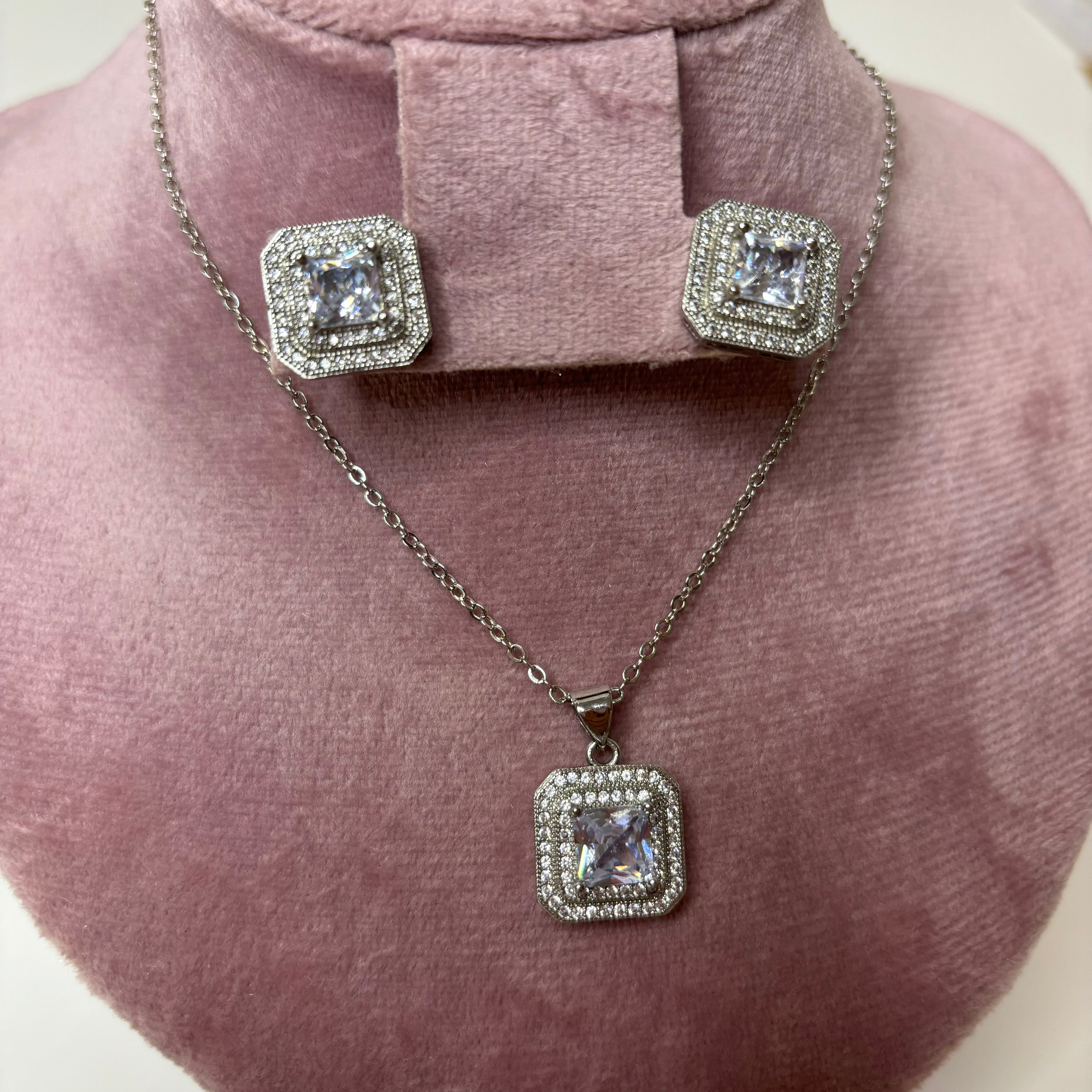A silver necklace with a square pendant and matching square stud earrings displayed on a pink fabric.