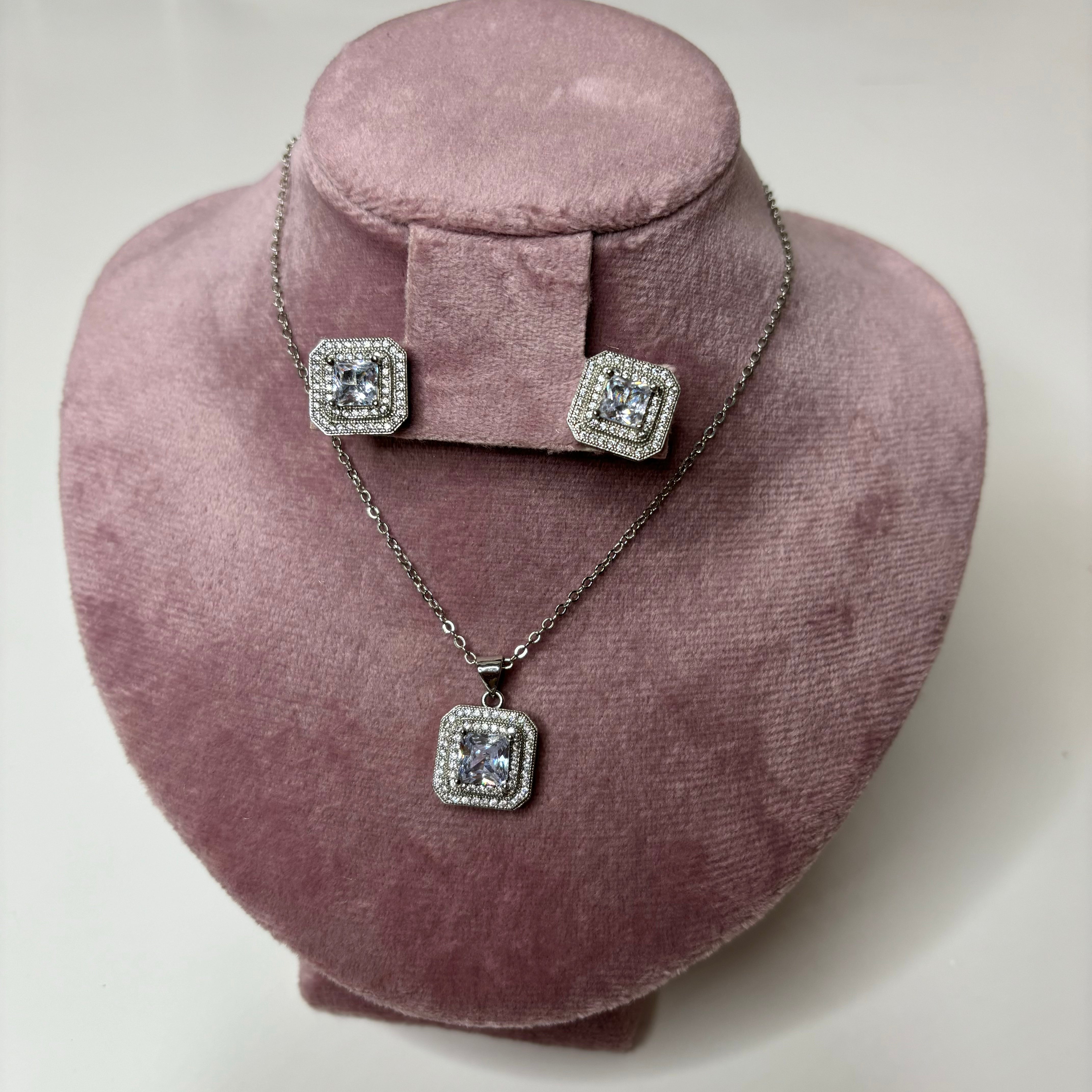 A silver necklace with a square pendant and matching square stud earrings displayed on a pink fabric.