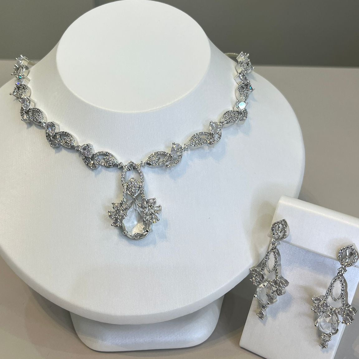 A luxurious silver necklace with a clear gemstone centerpiece and sparkling crystals, accompanied by matching earrings, displayed on a white bust.