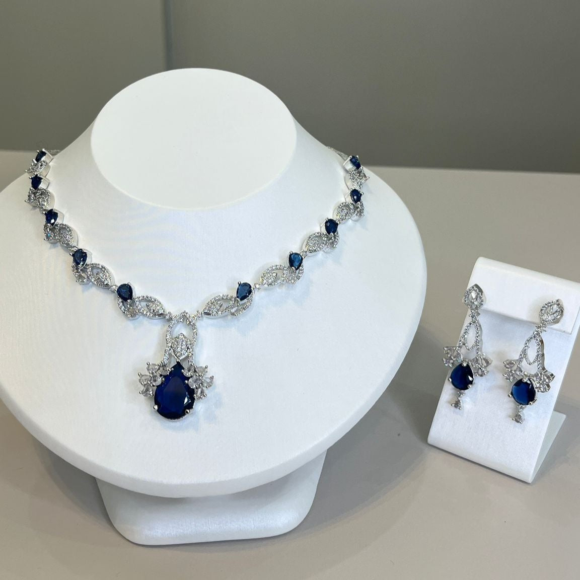 A luxurious silver necklace with a deep blue gemstone centerpiece and sparkling crystals, accompanied by matching earrings, displayed on a white bust.