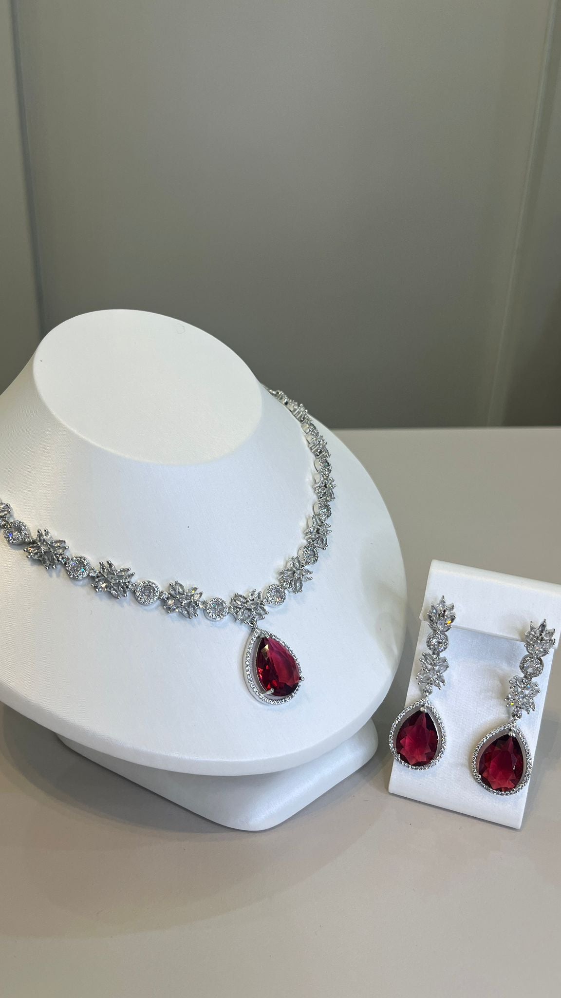 A silver-tone necklace with a large red teardrop gemstone centerpiece and sparkling crystal embellishments, accompanied by a pair of earrings that mirror the necklace's design, displayed on a white bust.