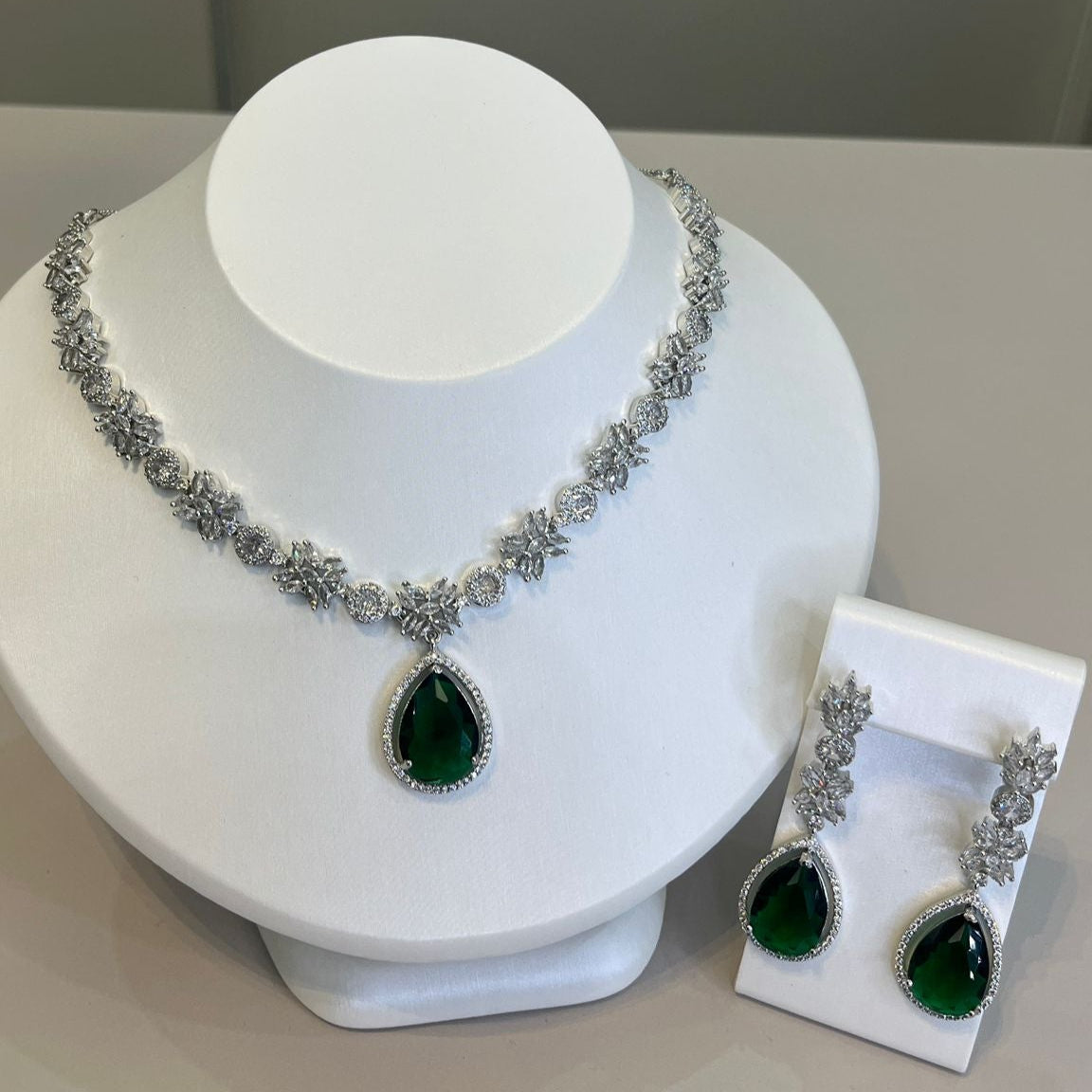 A silver-tone necklace with a large green teardrop gemstone centerpiece and sparkling crystal embellishments, accompanied by a pair of earrings that mirror the necklace's design, displayed on a white bust.