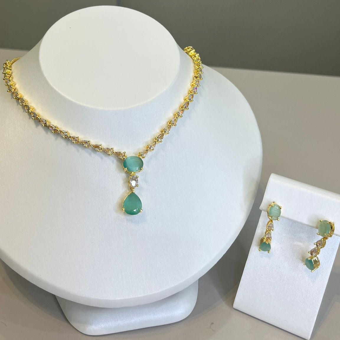 A teardrop necklace and earrings set displayed on a white mannequin. The set features a gold-tone chain with crystal embellishments and a teardrop pendant.