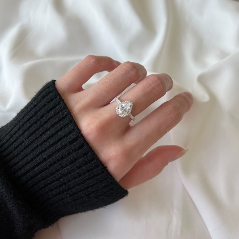 A sterling silver ring featuring a pear-shaped central stone with a halo of smaller stones around it, set on a white background on a women's hand.