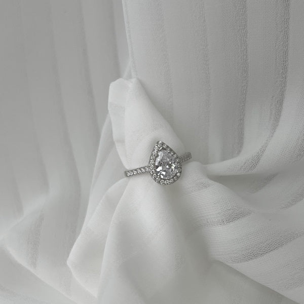 A sterling silver ring featuring a pear-shaped central stone with a halo of smaller stones around it, set on a white fabric in the background.