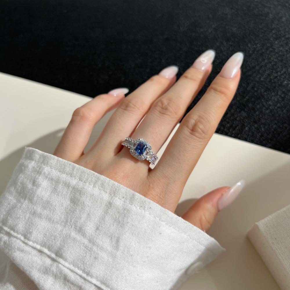A blue rectangular radiant cut CZ ring with a halo of smaller clear stones around it, displayed on a women's hand.