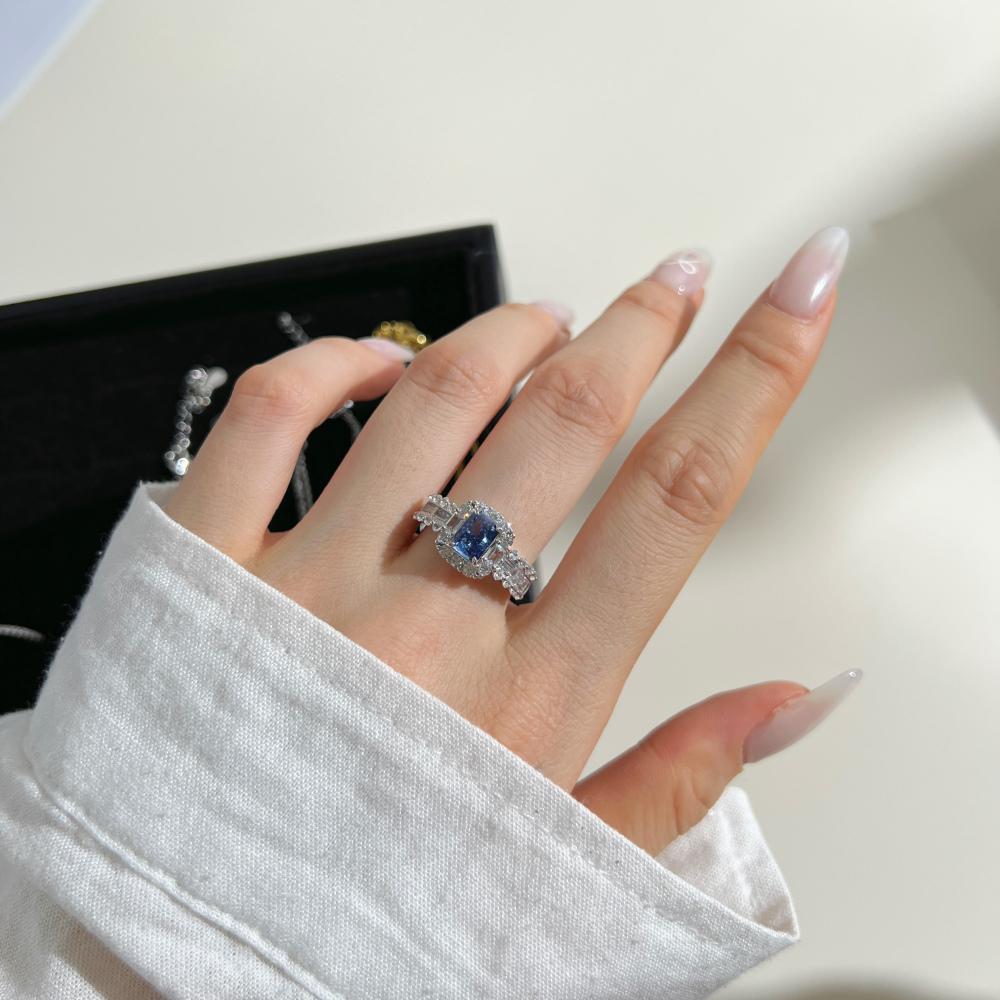 A blue rectangular radiant cut CZ ring with a halo of smaller clear stones around it, displayed on a women's hand.