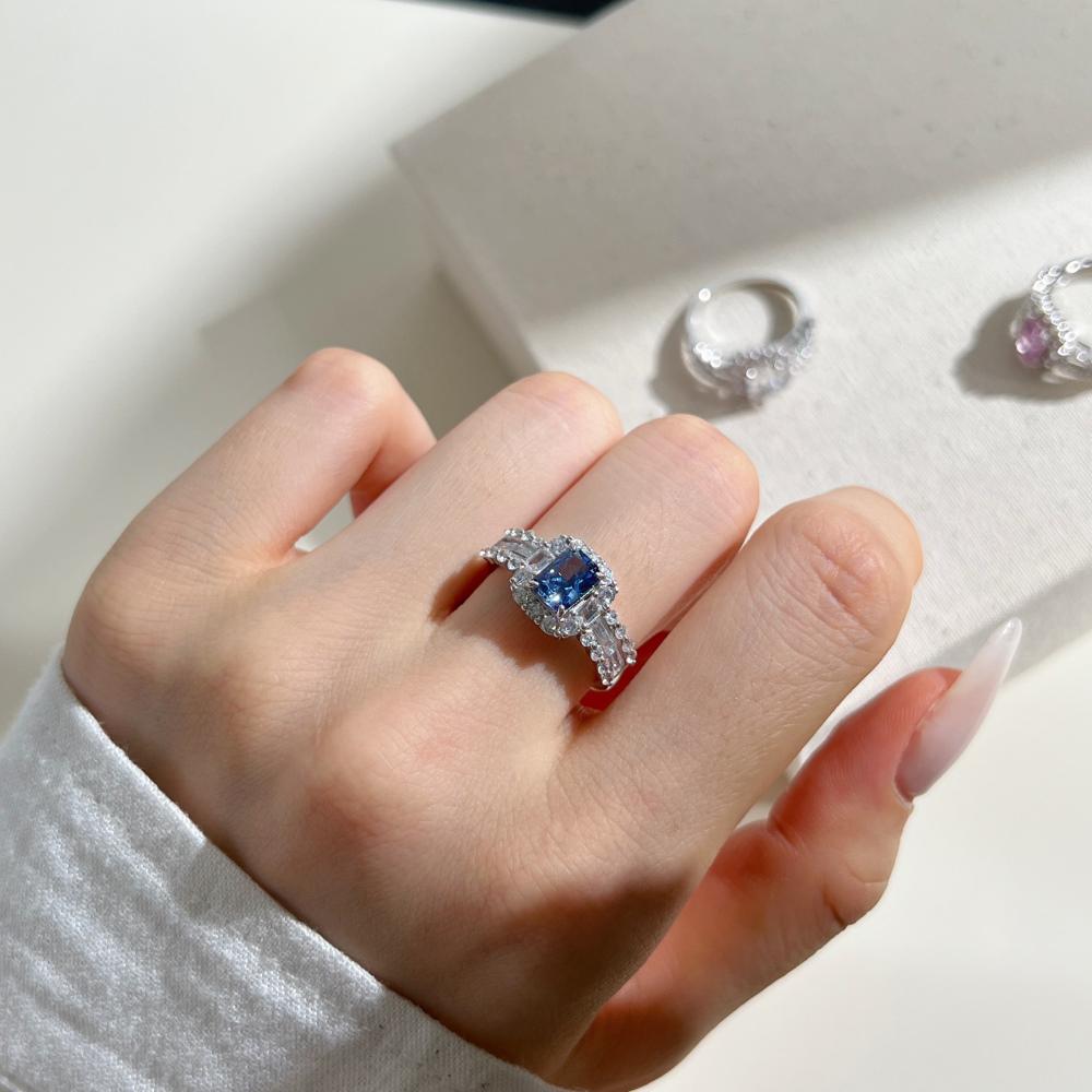 A blue rectangular radiant cut CZ ring with a halo of smaller clear stones around it, displayed on a women's hand.