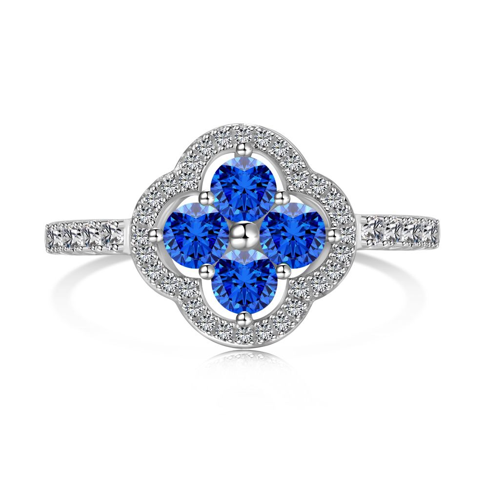 A clover-shaped ring with blue sapphire leaves and a round central sapphire, encircled by a halo of small clear stones, set against a white background.
