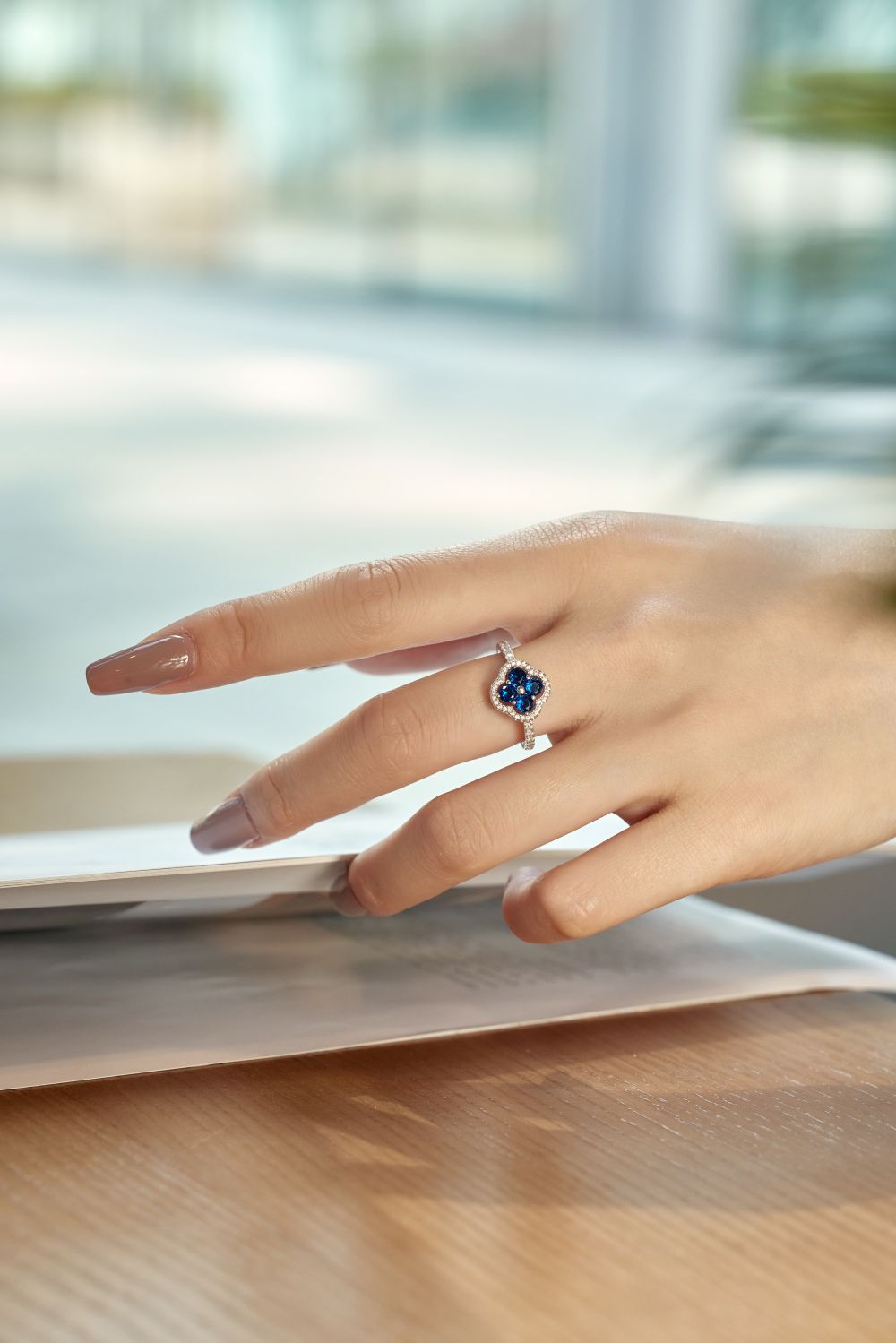 A clover-shaped ring with blue sapphire leaves and a round central sapphire, encircled by a halo of small clear stones, displayed on a women's hand.