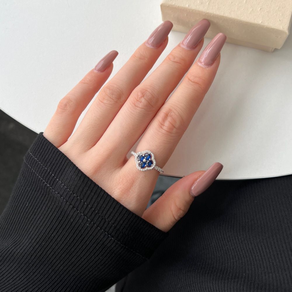 A clover-shaped ring with blue sapphire leaves and a round central sapphire, encircled by a halo of small clear stones, displayed on a women's hand.