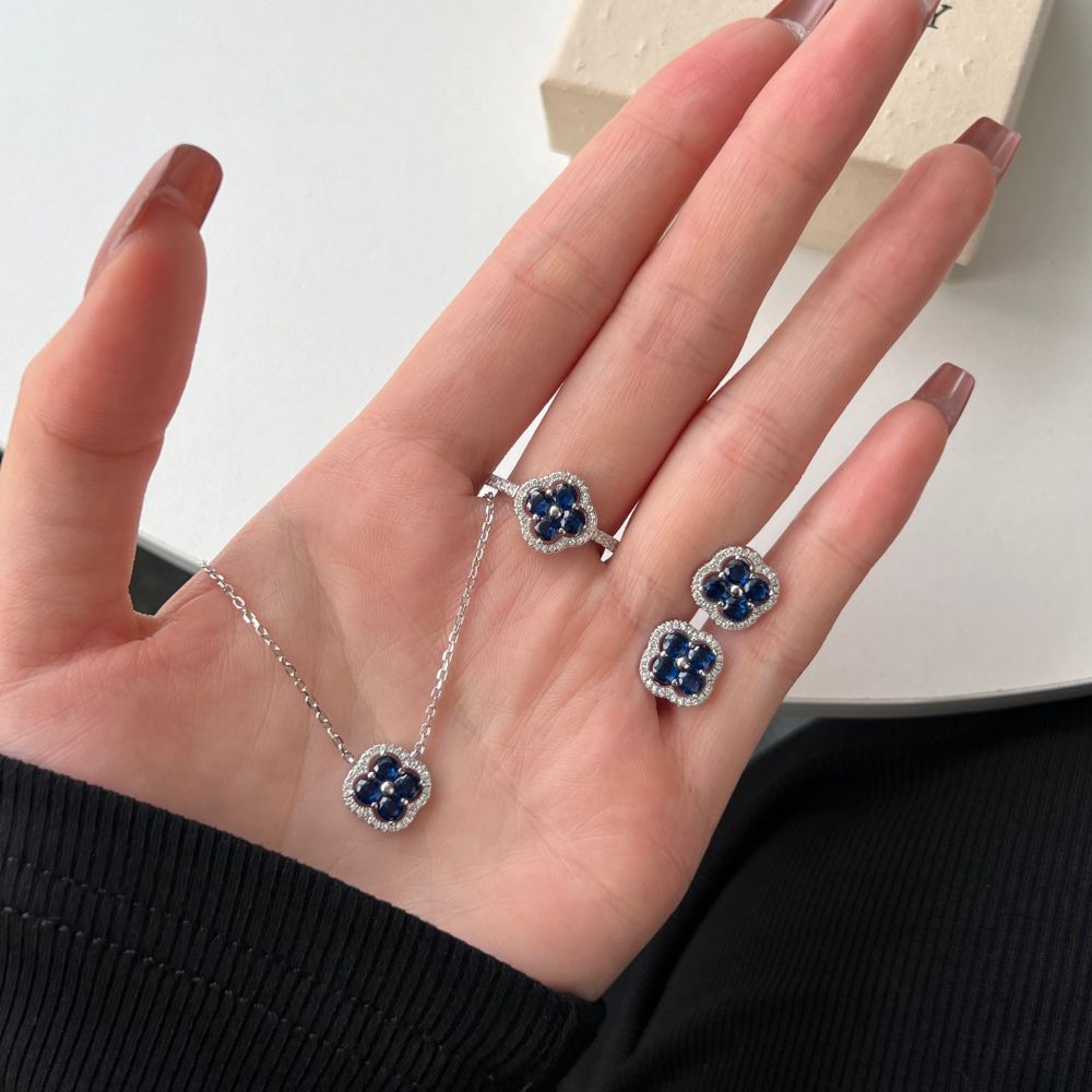 A silver necklace, earring and ring with a clover-shaped pendant adorned with blue cubic zirconia stones.