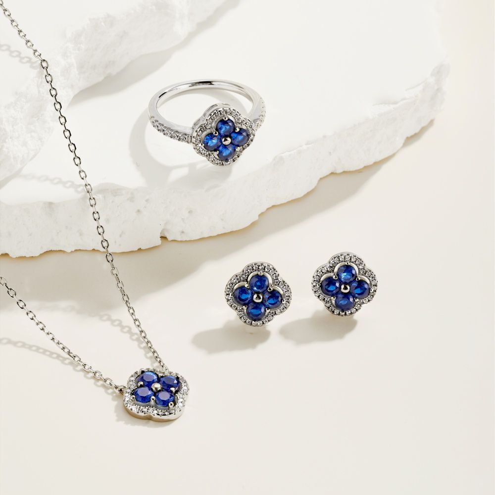 A silver necklace, earring and ring with a clover-shaped pendant adorned with blue cubic zirconia stones.