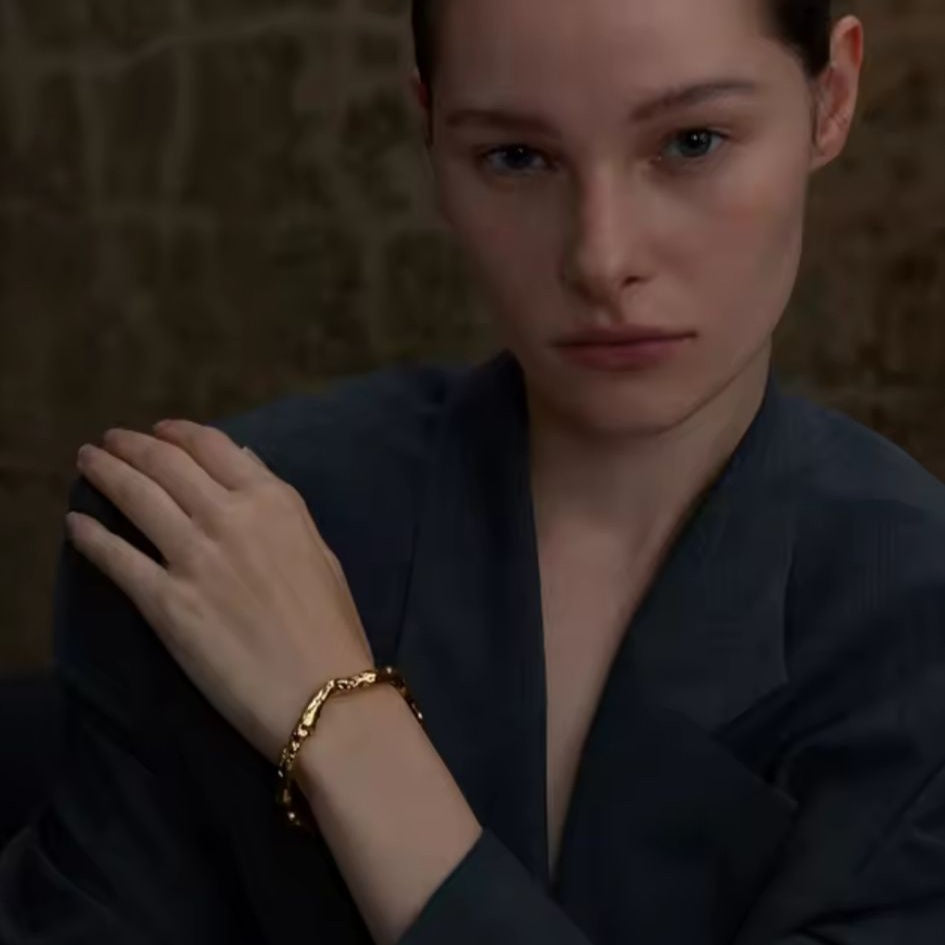 Woman wearing a dark outfit with a gold Hammered bracelet against a textured wall.
