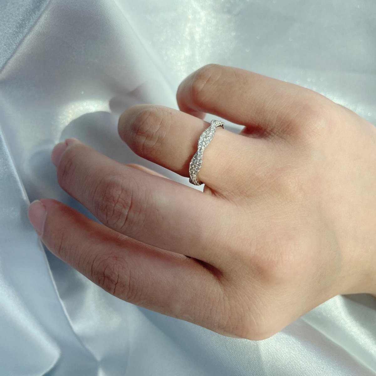 A sterling silver twist ring with multiple pave-set crystals adorning its surface on a female's hand on a white fabric in the background.