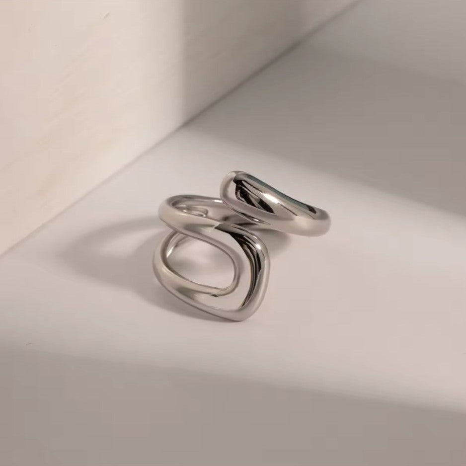 A silver-colored stainless steel hollow layered wide ring is displayed on a white surface in the background.
