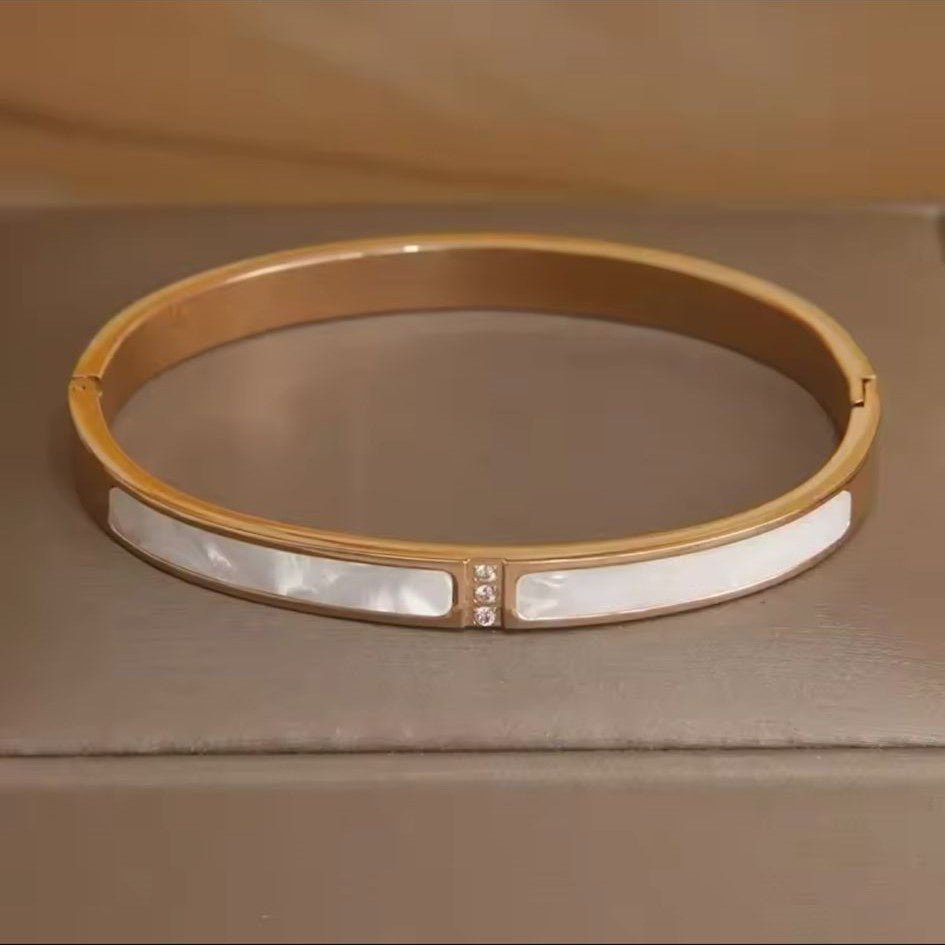 A gold stainless steel bangle bracelet with a pearl shell accent, displayed on a textured surface.