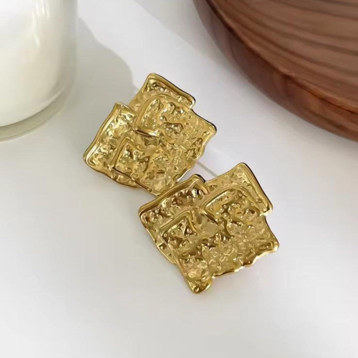 Gold square earrings on a white surface with a wooden object in the background
