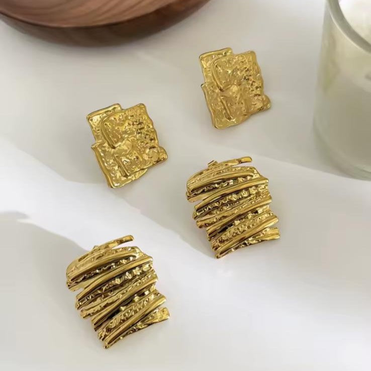Gold textured earrings on a white surface with a wooden bowl and glass in the background.
