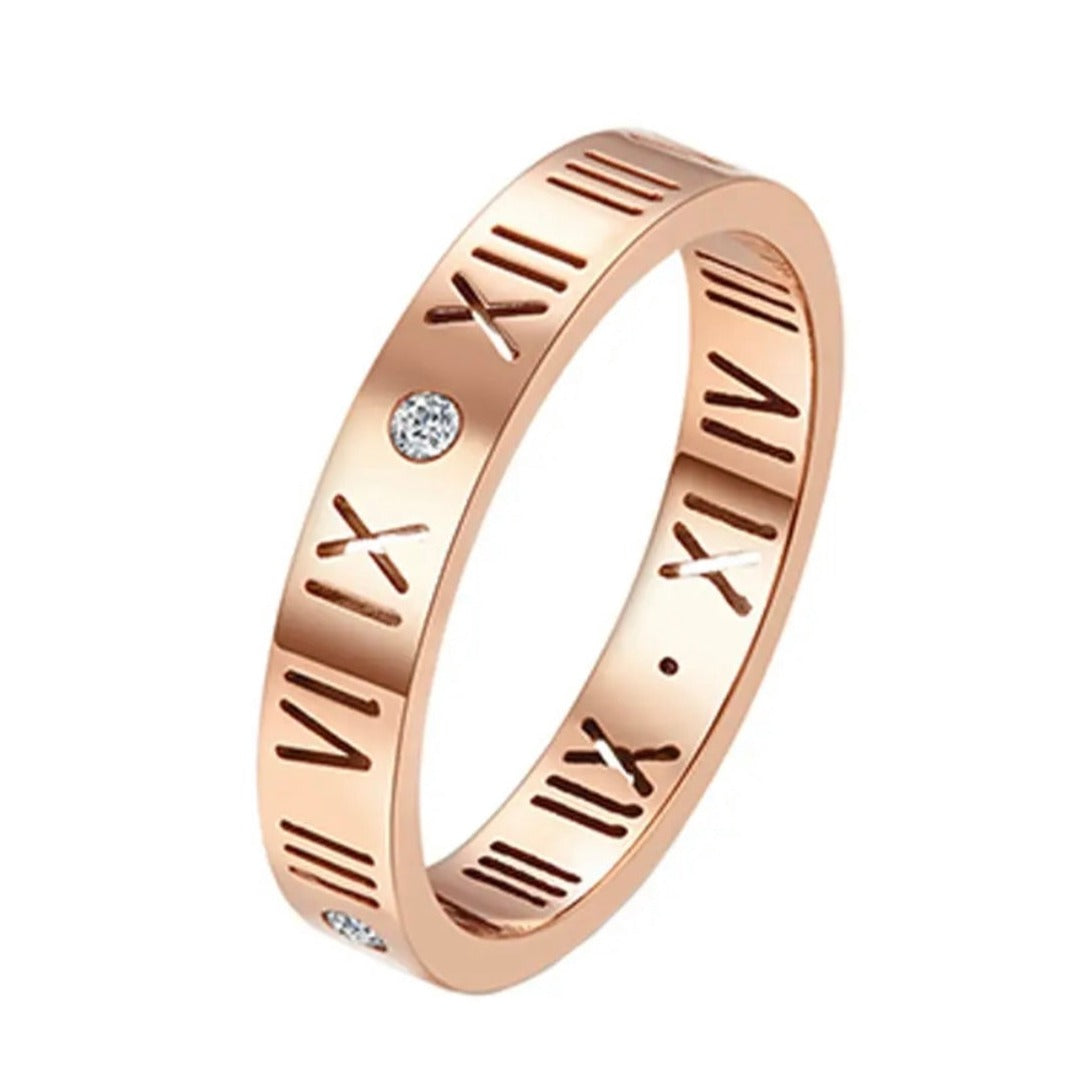 A gold-colored stainless steel band ring with Roman numeral patterns and small embellishments.