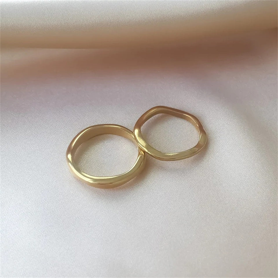 A gold-colored band ring with a wavy design, made from stainless steel, displayed on a textured surface.