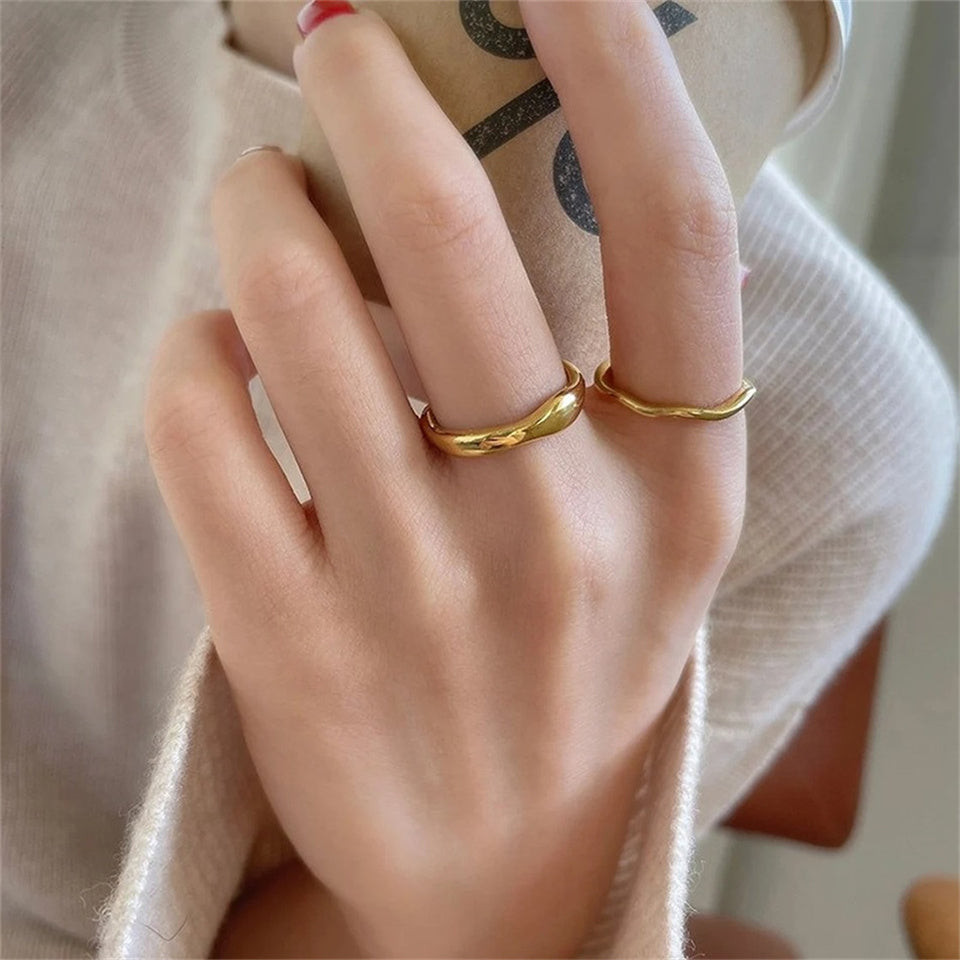 A gold-colored band ring with a wavy design, made from stainless steel, displayed on a female's hand.