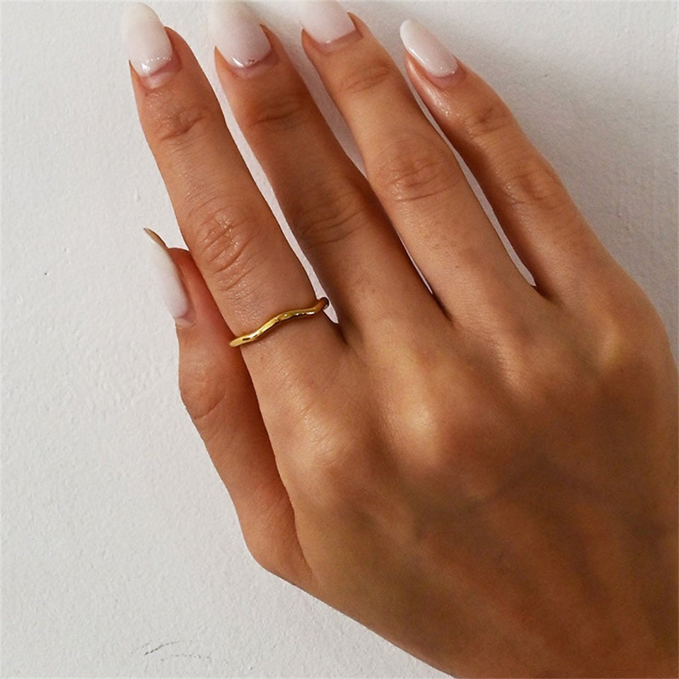 A gold-colored band ring with a wavy design, made from stainless steel, displayed on a hand in plain background.