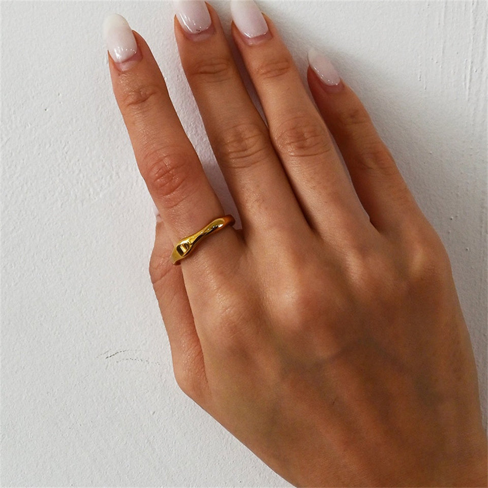 A gold-colored band ring with a wavy design, made from stainless steel, displayed on a female's hand on a plain background.