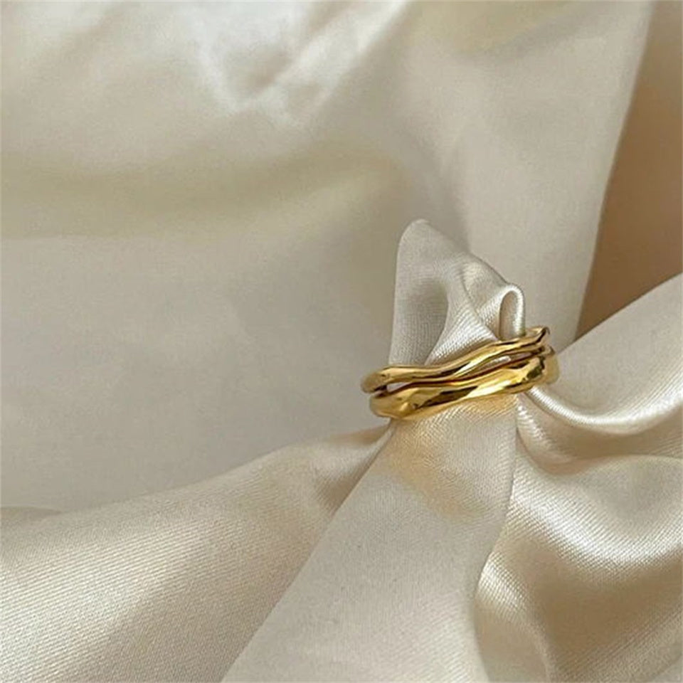 A gold-colored band ring with a wavy design, made from stainless steel, displayed on a white fabric in the background.