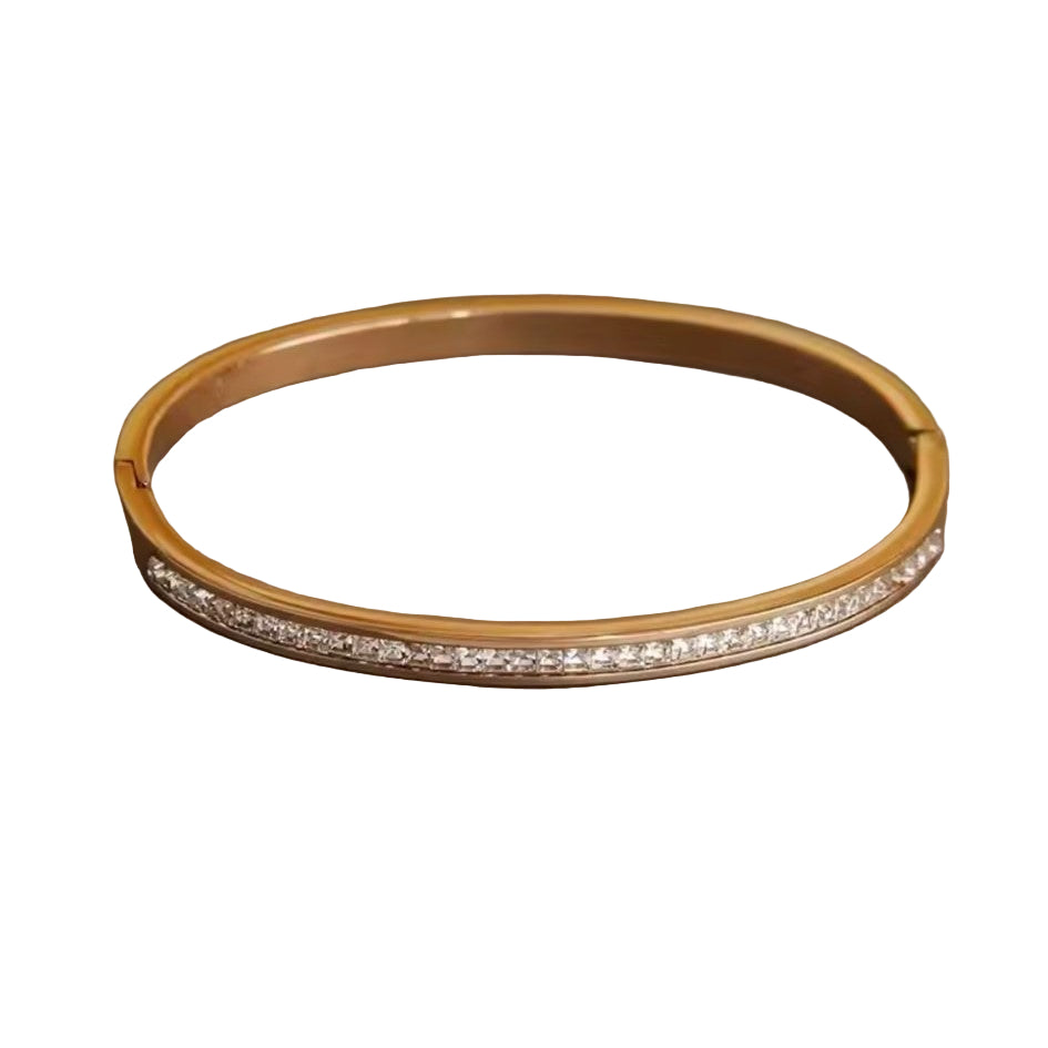 A gold stainless steel bangle bracelet with diamond accents displayed on a textured surface.