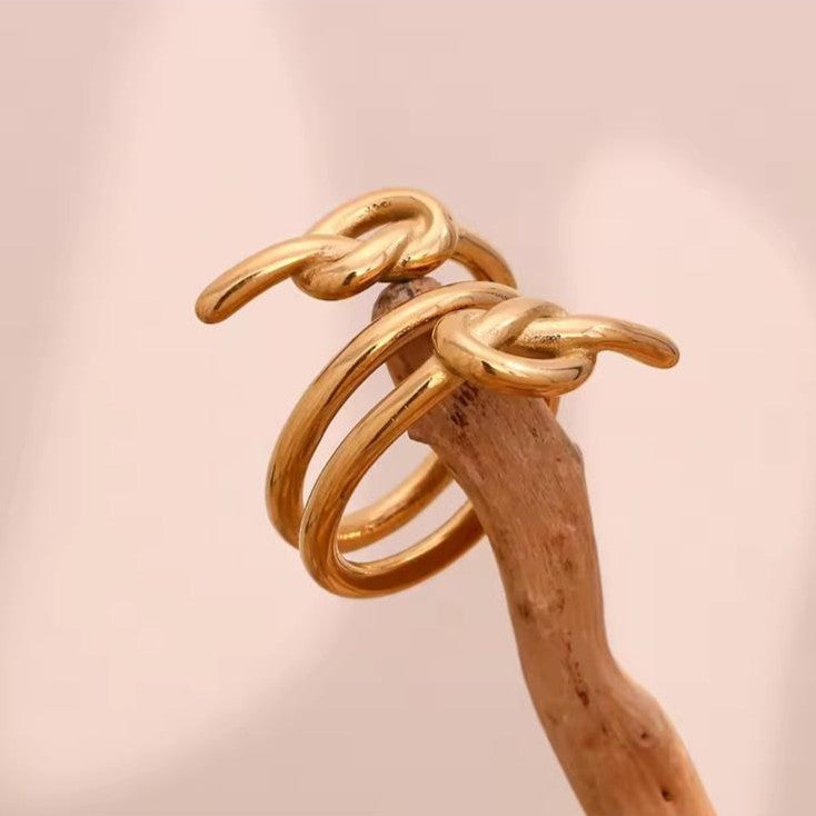 Gold spiral three-knot ring on a wooden stick against a pink background