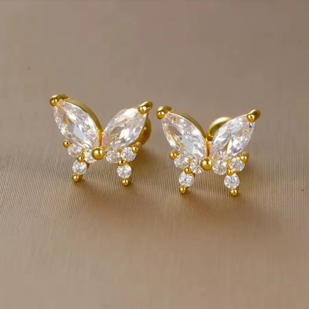 Butterfly-shaped earrings with gold and clear gemstones on a beige background