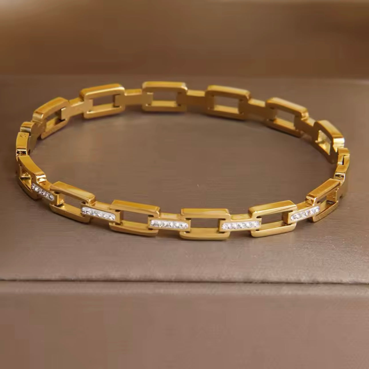A stainless steel hollow geometric bangle, with a gold finish, displayed on a textured fabric surface.