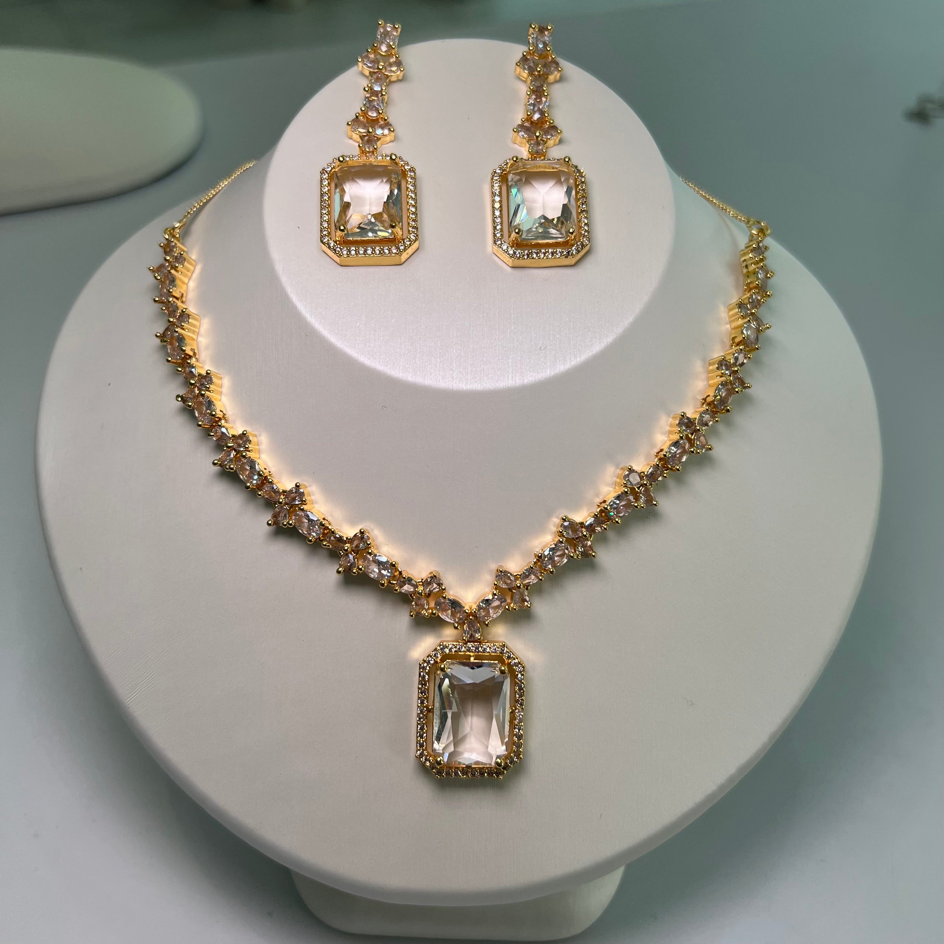 A gold-tone crystal charm necklace and earrings set with a rectangular centerpiece and drop earrings on a mannequin.