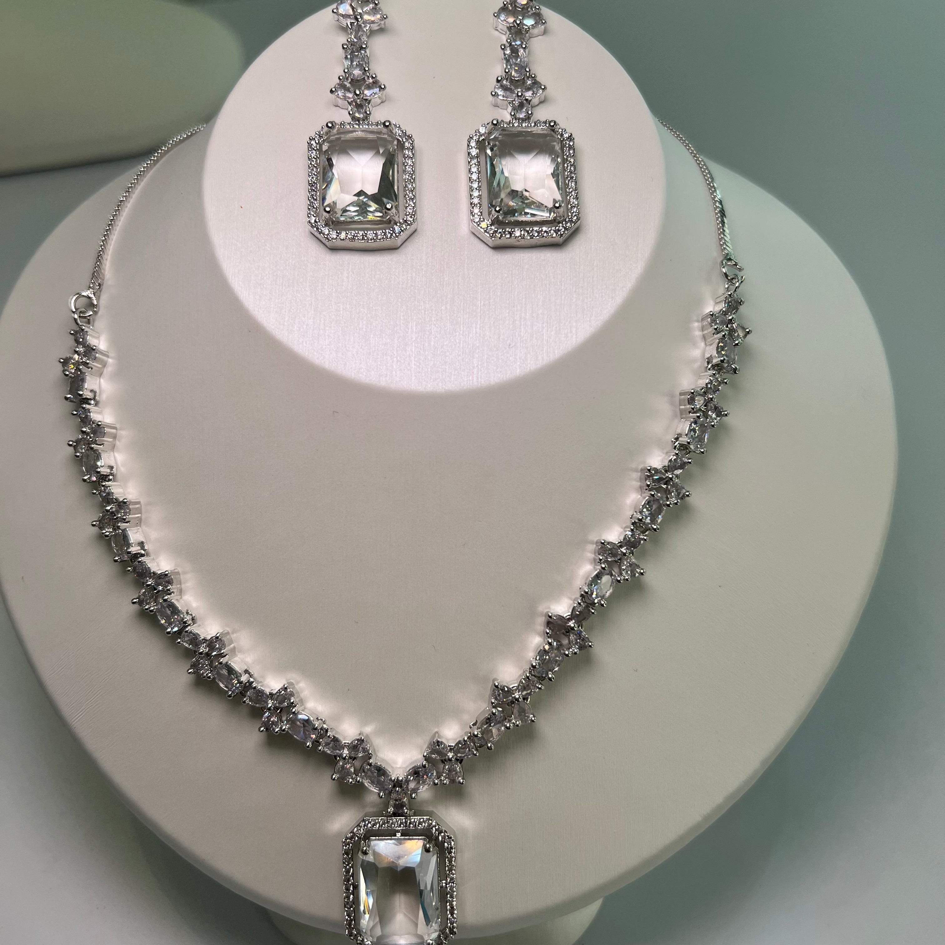 A silver-tone crystal charm necklace and earrings set with a rectangular centerpiece and drop earrings on a mannequin.